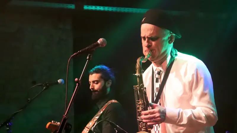 A saxophonist in a white shirt and checkered tie playing on stage next to a guitarist under green stage lights