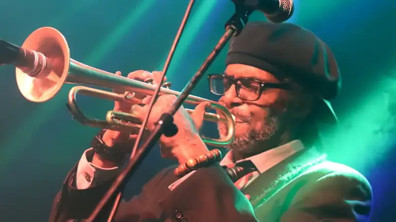 The band's trumpet player in a beret and glasses playing into a microphone under green and blue stage lights