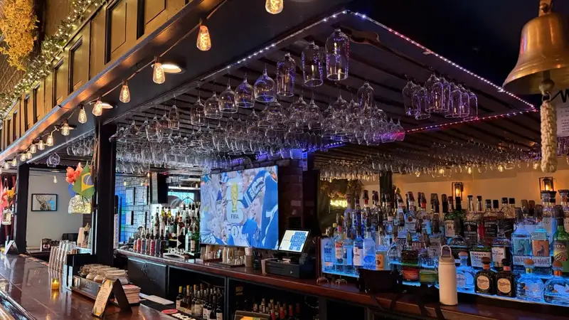 The long wooden bar at Off The Brook with hanging wine glasses, string lights, TVs and shelves of bottles