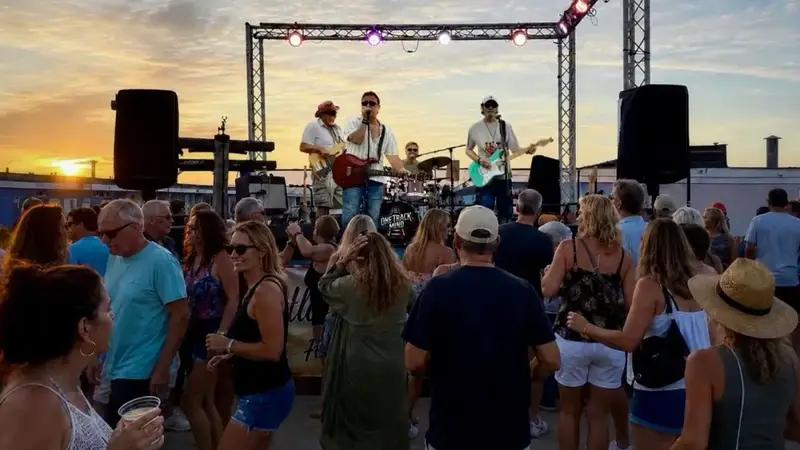 A crowd dancing on an outdoor deck at sunset while One Track Mind plays on a stage frame in the background