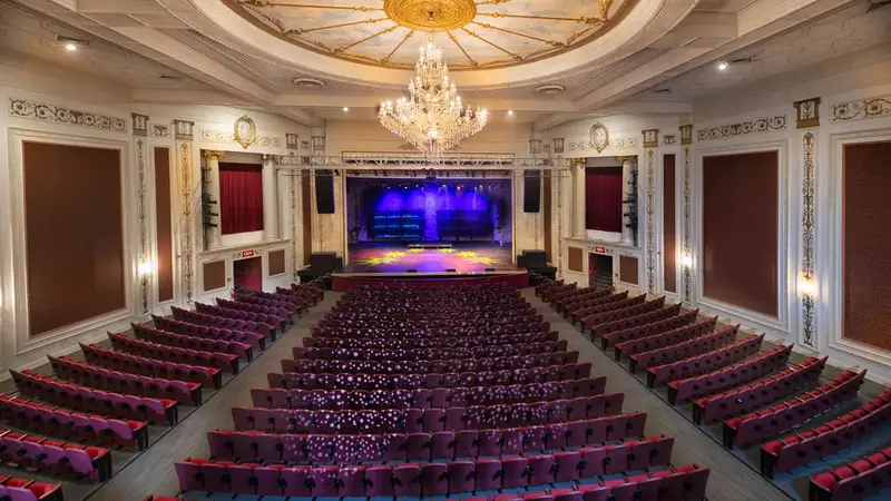 Inside the theater: rows of red seats facing a lit stage under a painted dome ceiling and a crystal chandelier