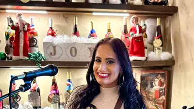 Renee D'Angelo smiling with her acoustic guitar at a microphone in a cozy room with wine bottles on shelves