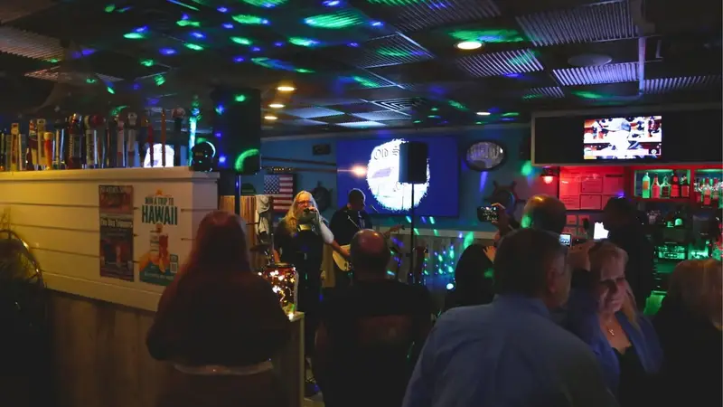 A crowd stands and watches a band at the far end of the room, with disco lights on the ceiling and TVs above the bar