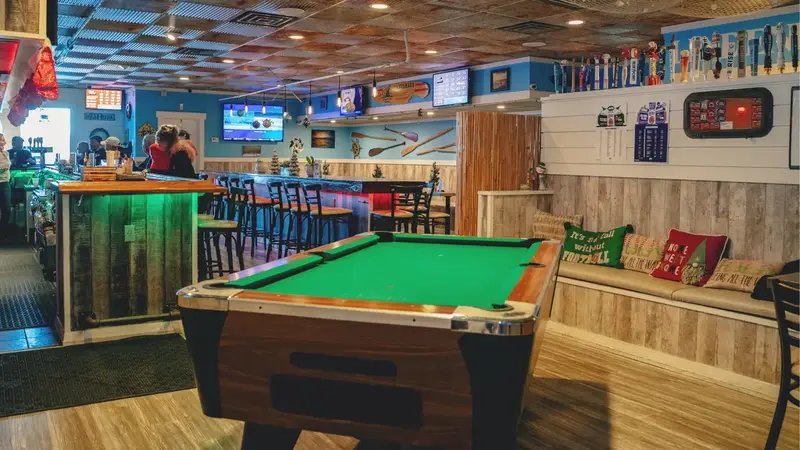 The bright bar room with a green pool table, a wooden bar with stools and beach-style wood paneling