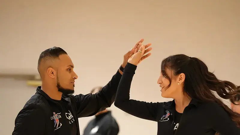 A man and a woman in matching black jackets practice a salsa turn, hands joined above their heads, at a dance festival class