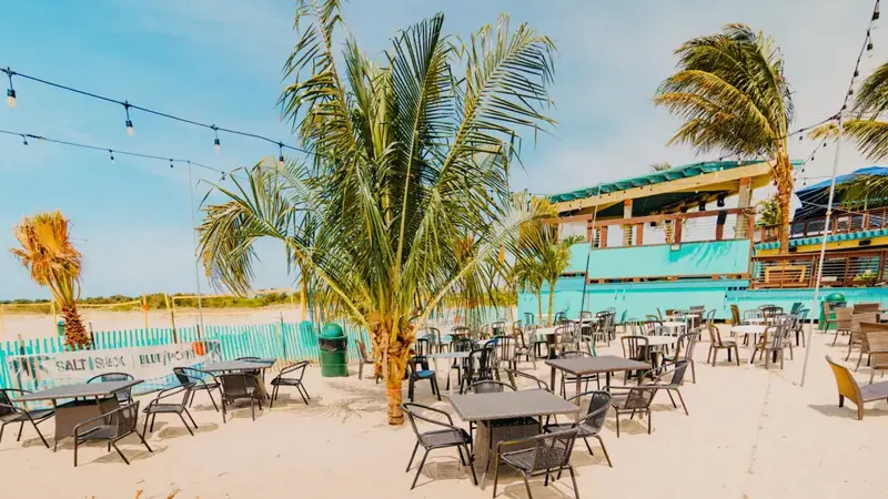 Tables and chairs set out on the sand under palm trees and string lights, in front of the teal Salt Shack building and its covered upper deck