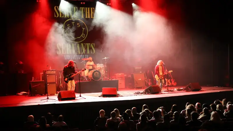 Serve The Servants playing a theater stage in red light and haze, with their band banner behind them and an audience in front