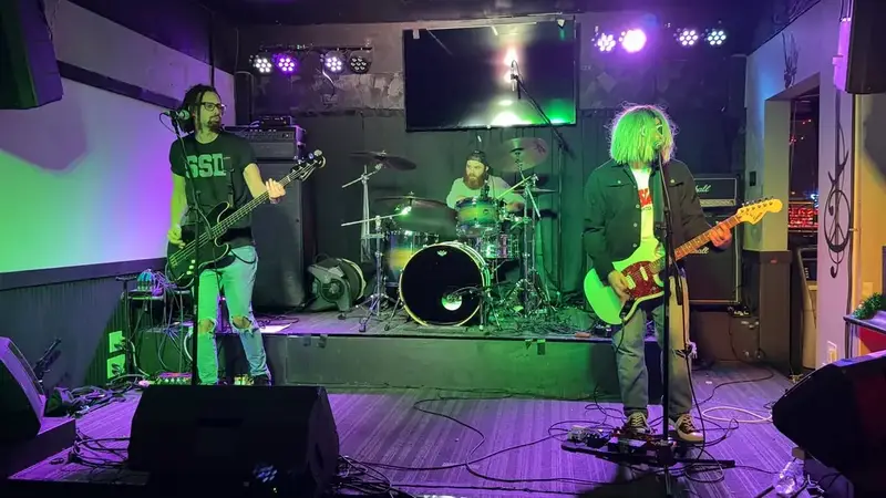 Serve The Servants on a small club stage under purple and green lights: bassist, drummer, and a singer-guitarist with green-blond hair