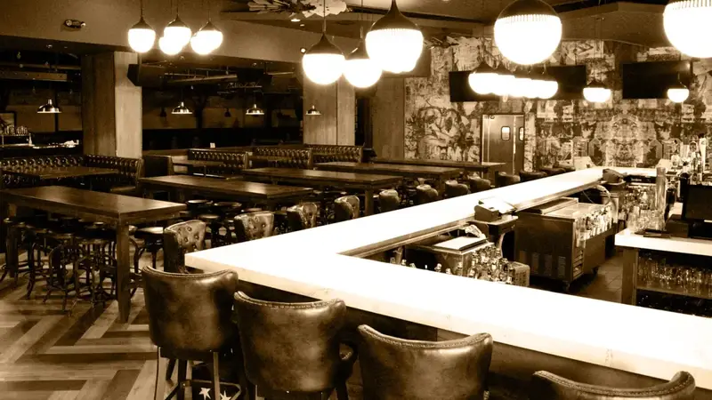 Sepia photo of a Stereo Garden bar with a white counter, leather bar stools, round hanging lamps and long tables