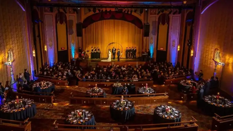 View from the balcony of the Suffolk Theater's Art Deco hall, with round dinner tables on tiered floors in front of the gold-curtained stage