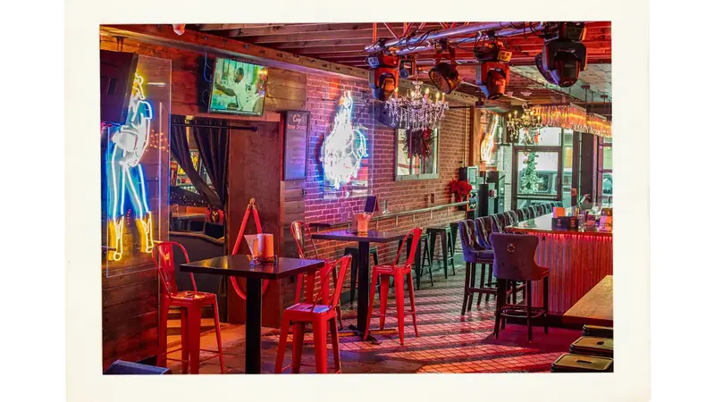 The bar room with red stools, high-top tables, neon signs and chandeliers over a striped wood floor