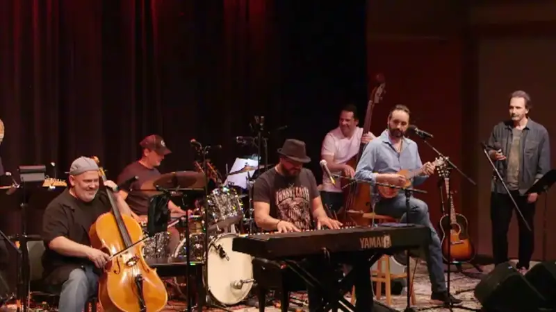 Band members playing cello, piano, upright bass, drums and guitars on a stage with red curtains