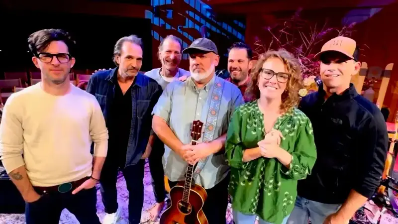 Seven musicians of The Cosmic Orchestra standing together on a stage, one holding an acoustic guitar