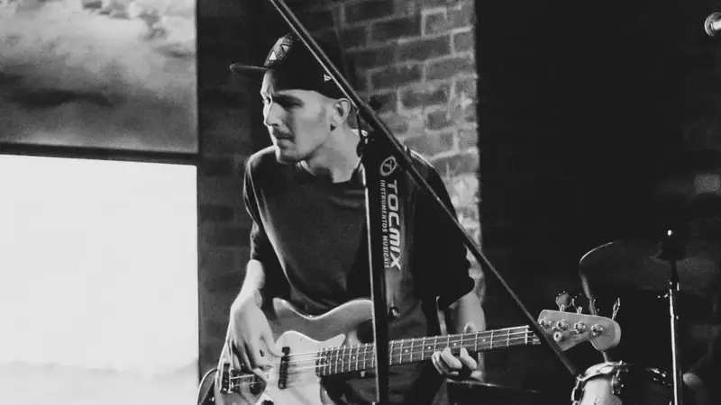 Black-and-white photo of the band's bass player in a backwards cap playing on stage next to a drum kit