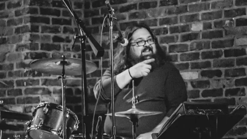 Black-and-white photo of the band's drummer smiling behind his drum kit in front of a brick wall