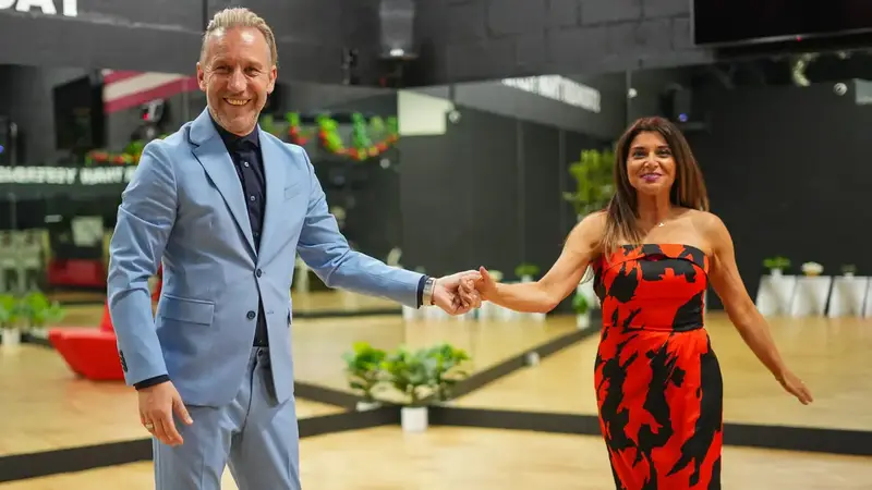 A man in a light blue suit and a woman in a red and black dress dancing hand in hand on the studio floor