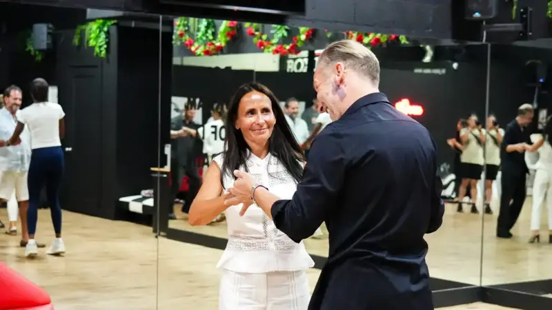 A teacher leading a student in a Latin dance step in front of the studio's mirror wall while other students practice nearby