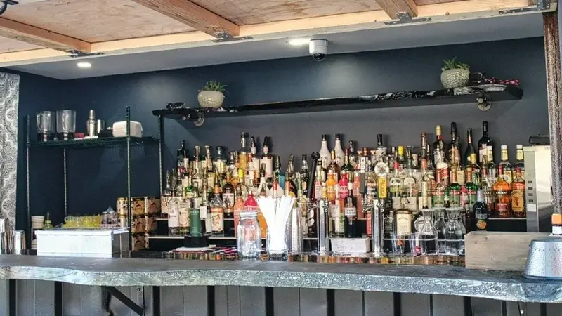 A bar counter with rows of liquor bottles on shelves against a dark blue wall under a wood-beam ceiling