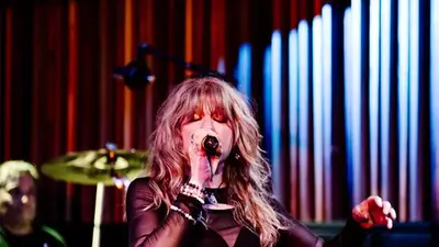 A singer with long blond hair sings into a microphone on stage, with a drummer behind her and colored stage lights