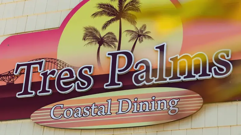 The Tres Palms Coastal Dining sign with palm trees on the restaurant's shingled outside wall