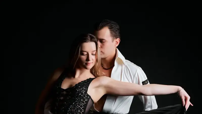Val, in a white dance shirt, holds Neda as she sweeps out a long black pleated skirt against a dark background