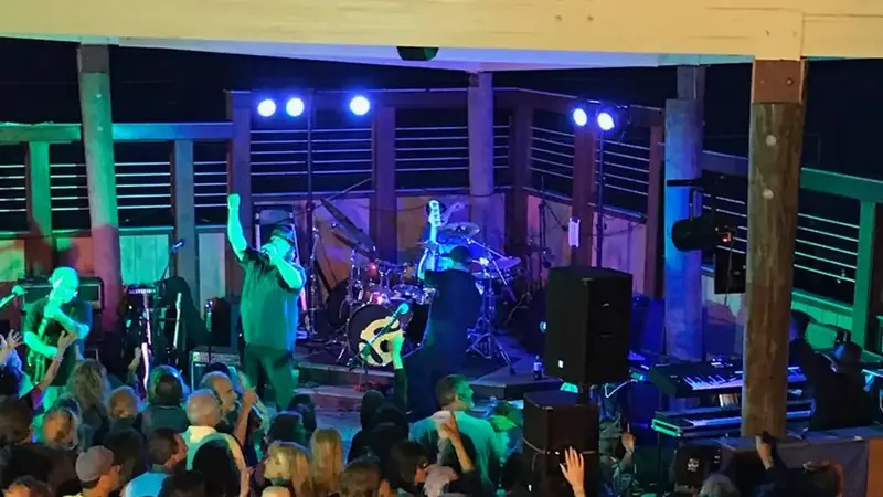 A packed crowd dancing with arms in the air in front of the band playing on an outdoor deck stage at night