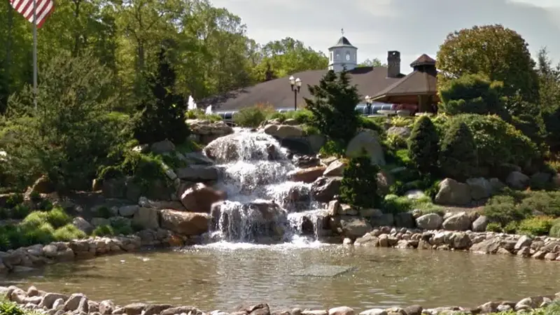 A stone waterfall and pond at The Waterfalls entrance, with the clubhouse and its cupola behind the trees