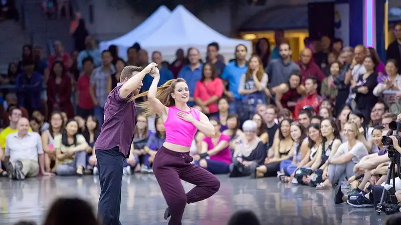 A couple dances a West Coast Swing demonstration on a shiny floor while a large crowd sits around them watching