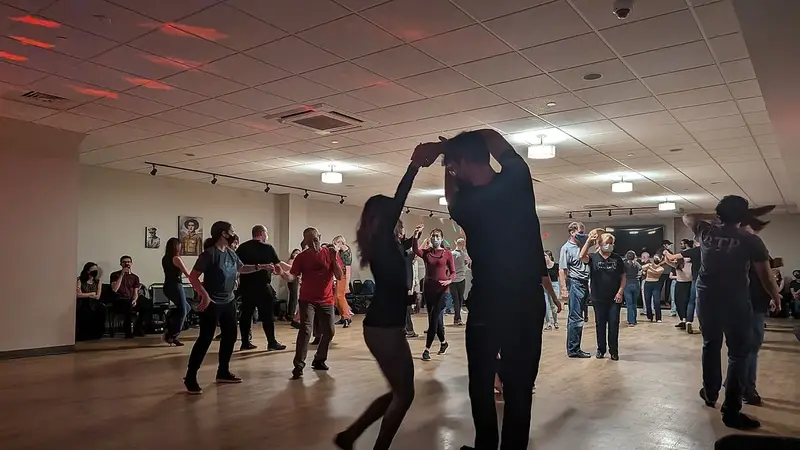 Couples dance West Coast Swing at a social dance in a low-lit community hall; in front, a man turns his partner under his arm